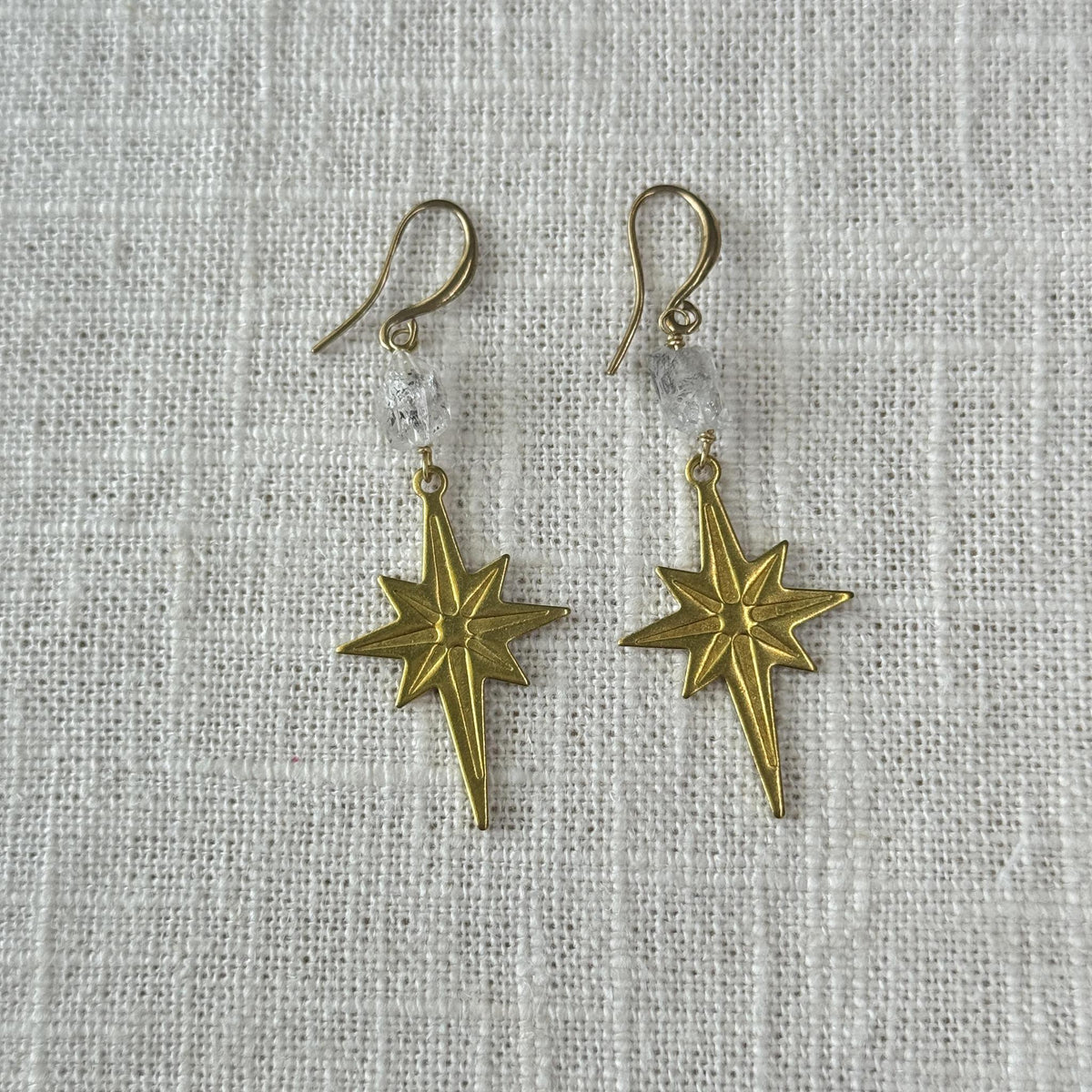 Spellcrafter’s Herkimer Diamond North Star Earrings feature gold star-shaped dangles and clear beads, ideal for meditation or balancing your Crown chakra. Displayed on textured white fabric, they bring subtle elegance to any look.