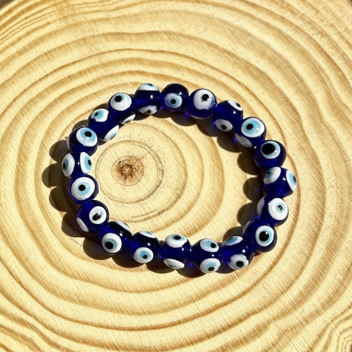 The Spellcrafter Evil Eye Beaded Bracelet, featuring dark blue beads with signature eye-like designs, is displayed in a circle on a wood surface with circular grain patterns to offer protection against negative energy.