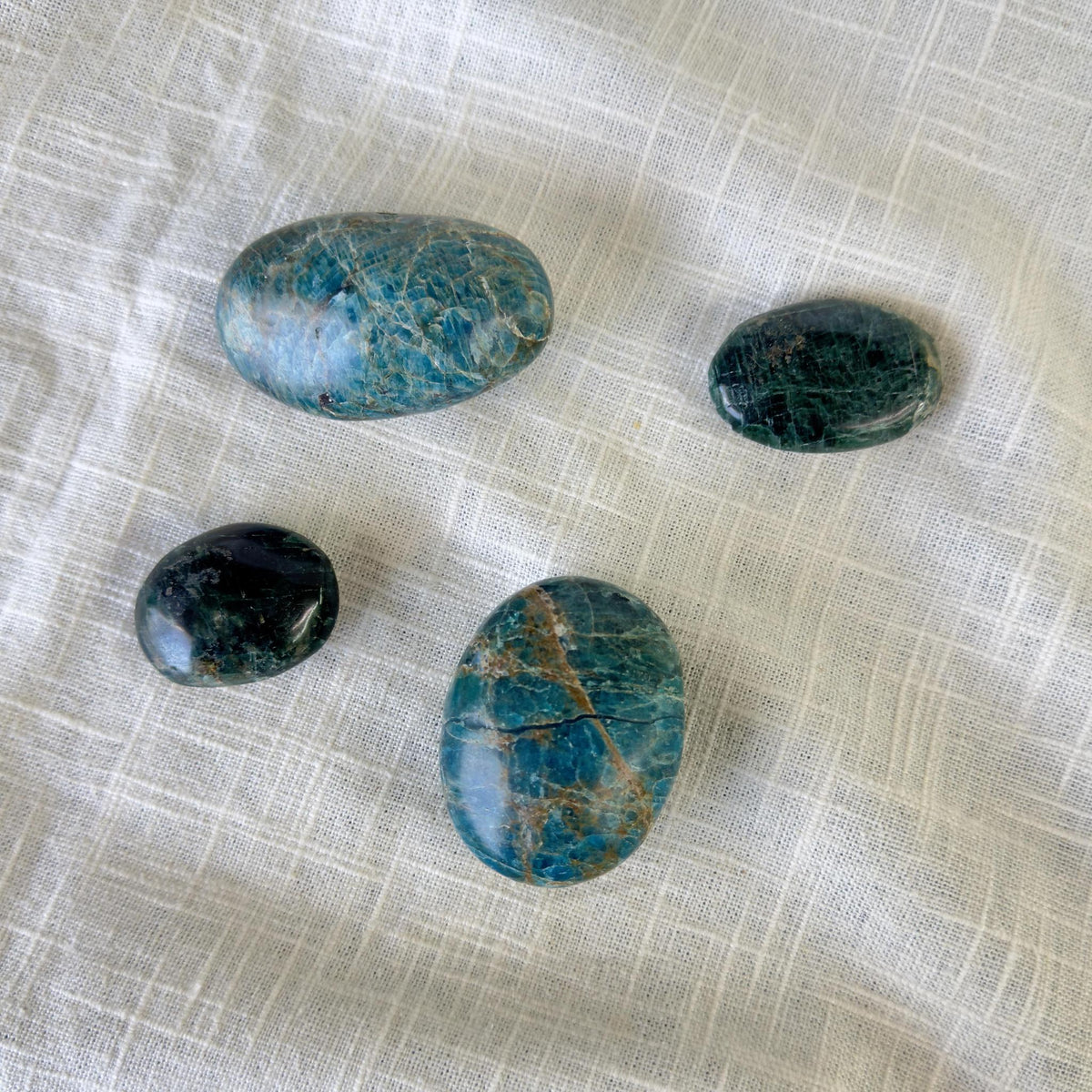 Four glossy Blue Apatite Palm Stones by Spellcrafter, featuring natural veining and varying sizes, rest on a textured white cloth. Associated with the Third Eye chakra, these polished gems are believed to enhance motivation.