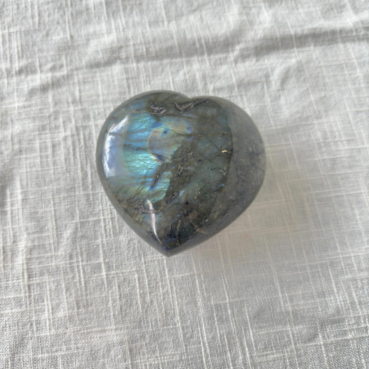 The Spellcrafter Labradorite Heart, with blue and green iridescent flashes, rests on textured white fabric, radiating creative energy and supporting chakra balance.
