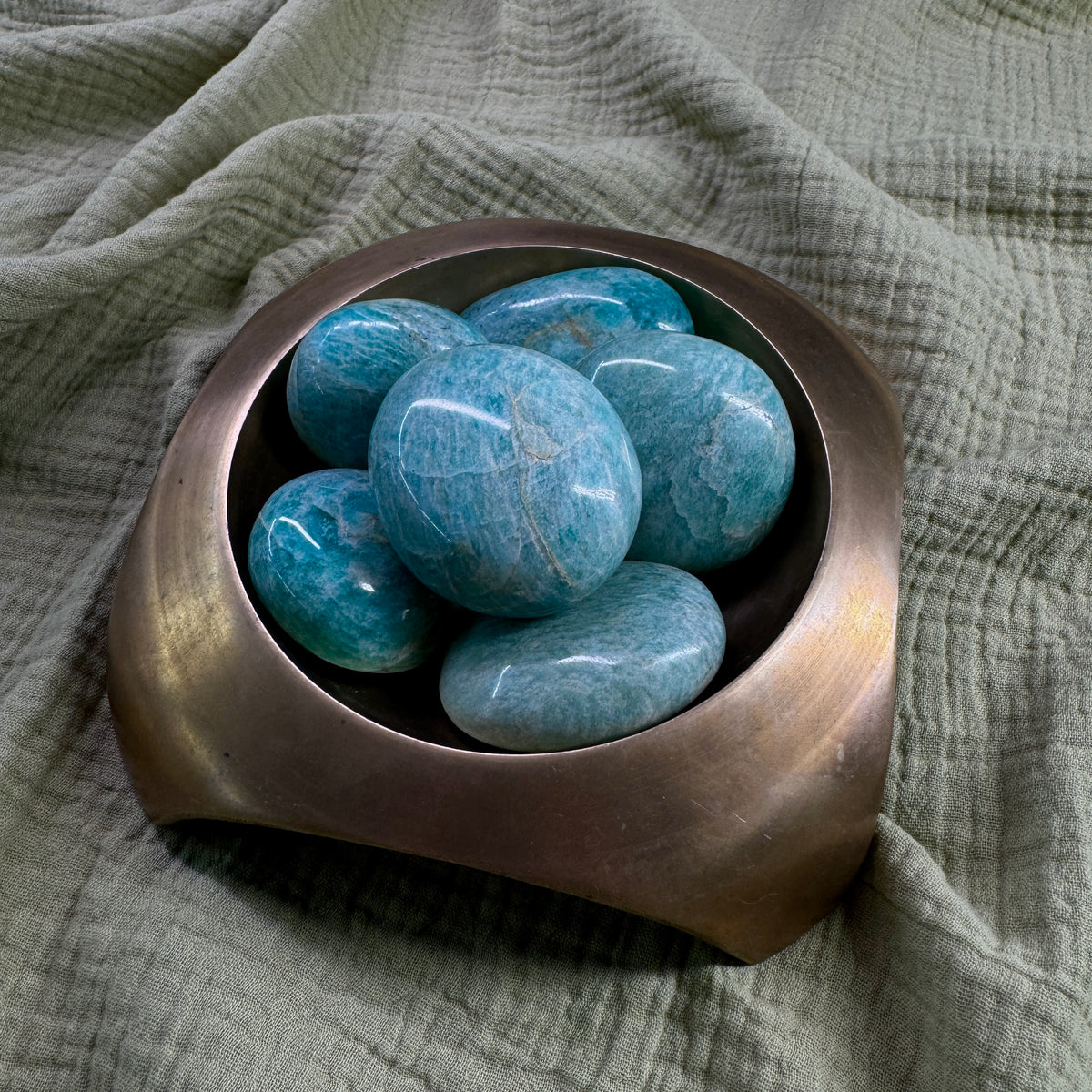 A bronze bowl displays several Spellcrafter Amazonite Palm Stones on a textured light green fabric background.