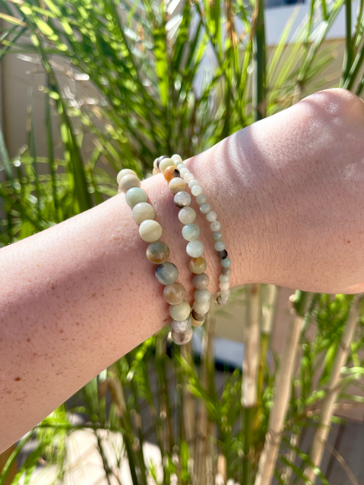A hand adorned with three Spellcrafter Sunstone Crystal Bracelets glimmers in sunlight, surrounded by tall green grass and a soft-focus pale fence in the background.