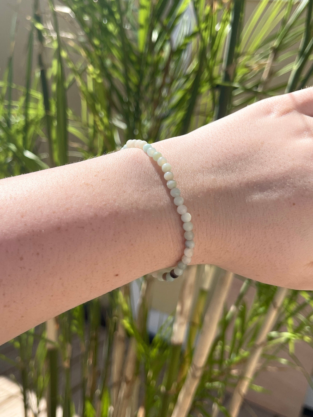 A close-up of a wrist wearing the Spellcrafter Sunstone Crystal Bracelet, featuring small round beads in pale blue, white, and light green for healing energy, with blurred green leafy plants in the background.
