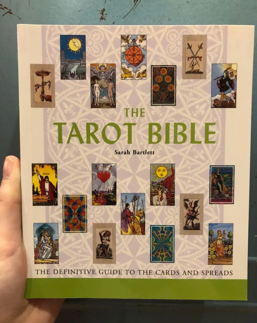 A hand holds Spellcrafter's The Tarot Bible. Its cover displays tarot cards and the subtitle "The Definitive Guide to the Cards and Spreads" on a pale patterned background.