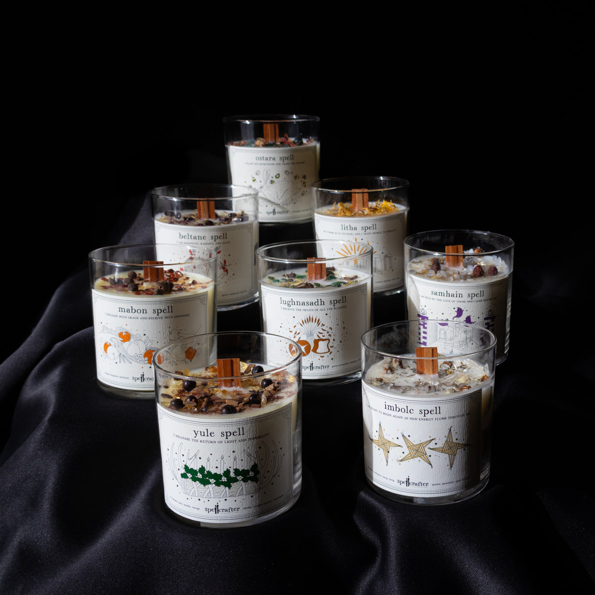 The Spellcrafter Wheel of the Year Candle Collection features eight decorated glass candles with wooden wicks, herbs, and crystals, arranged on silky black fabric—perfect for celebrating the seasons.