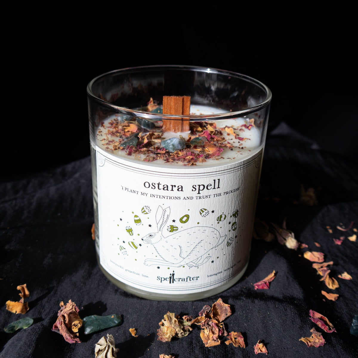 The Spellcrafter Ostara Spell Candle features a glass jar with a rabbit design, topped with dried flowers, herbs, and cinnamon stick. Infused with citrus essential oils, it rests on a dark surface among petals and stones.