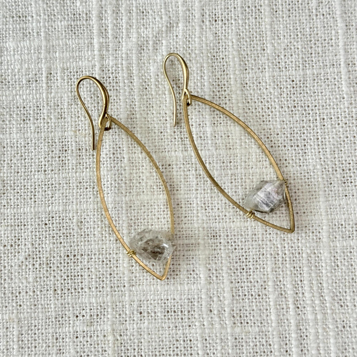 Spellcrafter's Herkimer Floating Marquise Earrings feature gold wire marquise shapes with a small, rough Herkimer Diamond at the base, shown on white textured fabric.