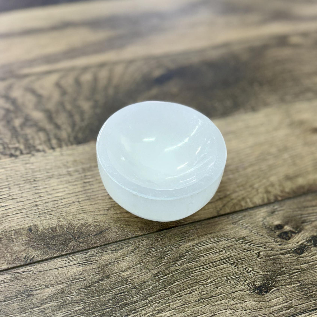 The Spellcrafter Selenite Round Mini Bowl, a translucent white piece, rests on a textured wooden surface, evoking cleansing and harmony.