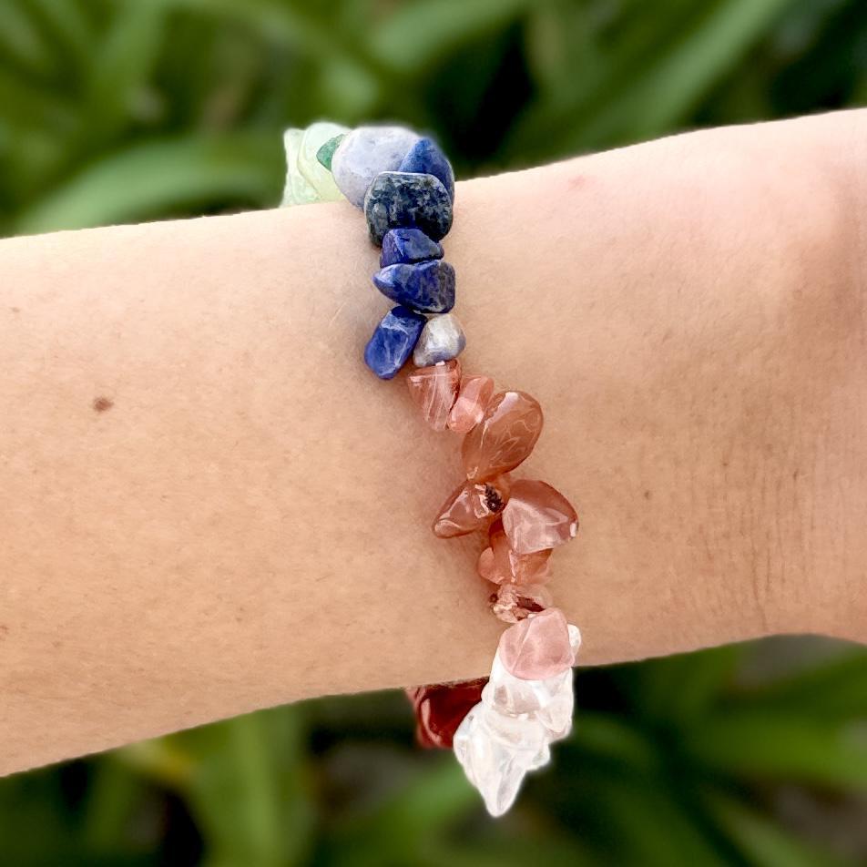 A close-up of a wrist wearing the Spellcrafter Chakra Stone Bracelet, showcasing vibrant blue, green, red, and clear gemstone chips against a softly blurred green background.