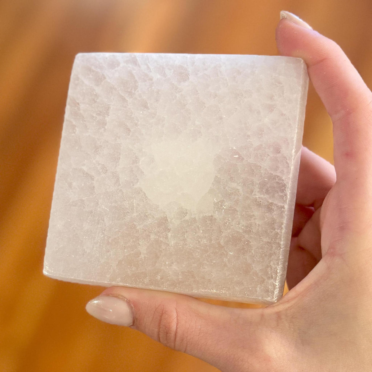 A hand with manicured nails holds the Spellcrafter Selenite Square Plate, its translucent, rough texture radiating harmony against a blurred warm-toned background.