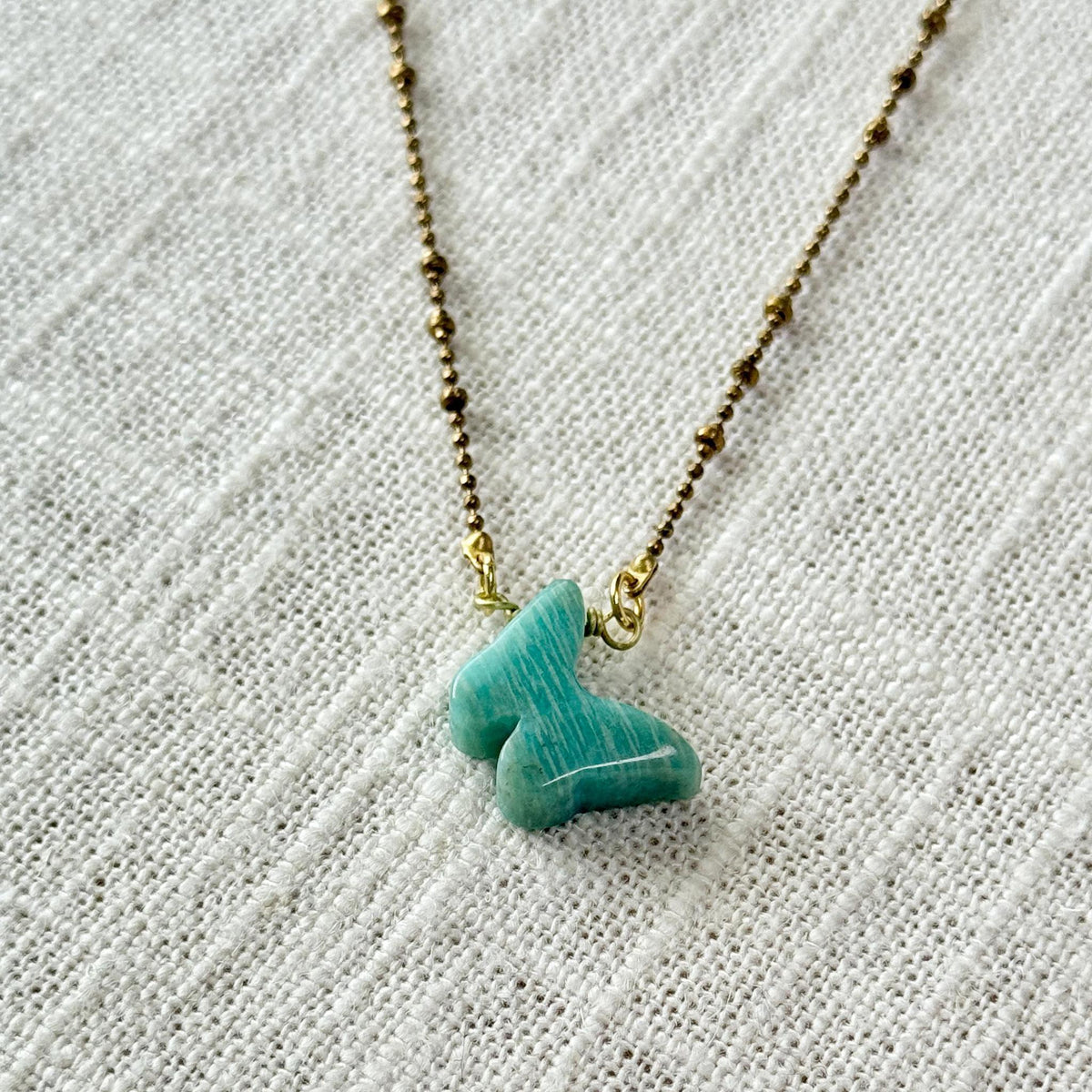 The Spellcrafter Amazonite Butterfly Necklace, featuring a gold beaded chain and turquoise butterfly-shaped pendant, is shown on a textured off-white fabric background.