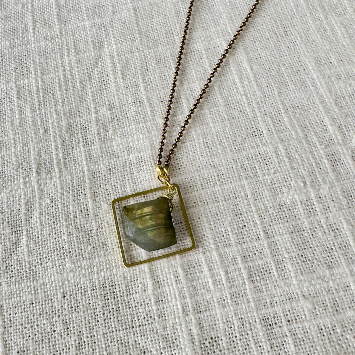 The Spellcrafter Labradorite Framed Diamond Necklace features a square, gold-framed greenish-gray labradorite pendant on a fine chain, displayed on light textured fabric—an elegant chakra jewelry piece for subtle energy and style.