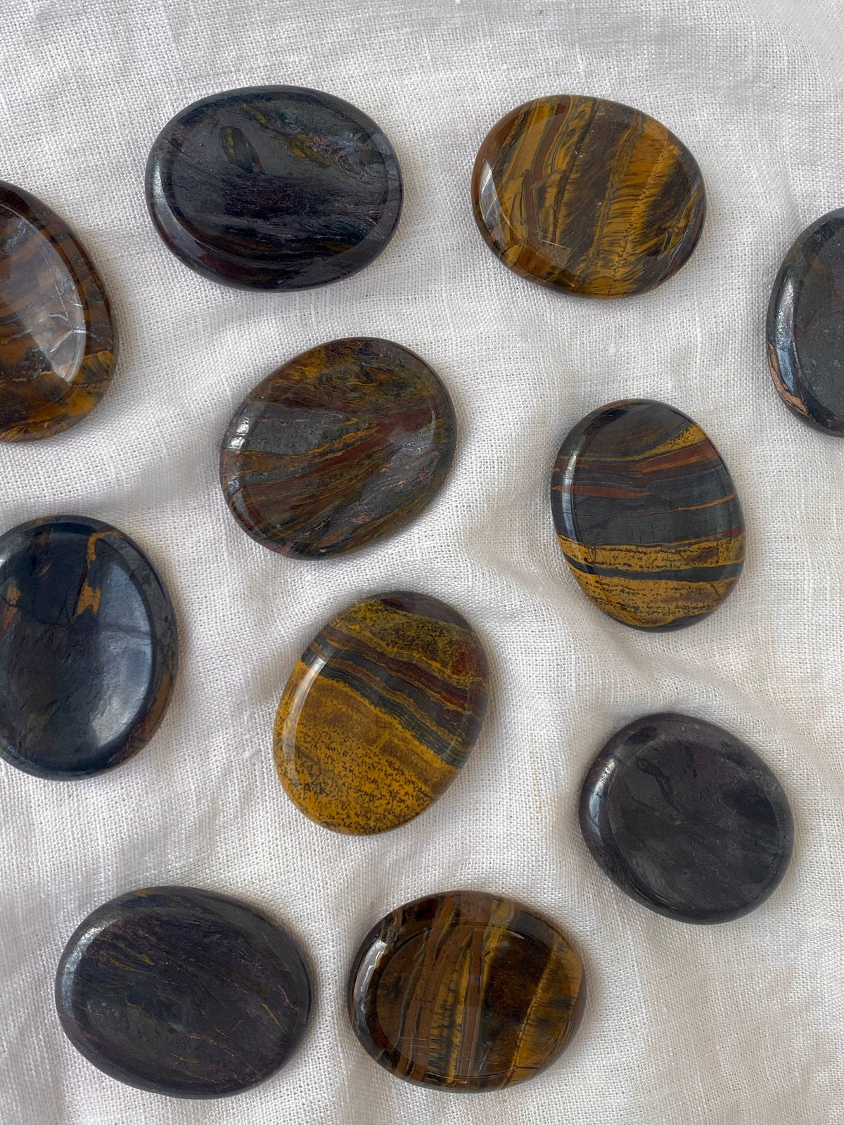 Spellcrafter’s Tiger Iron Calming Stones, featuring brown, gold, and black swirls, are arranged on white textured fabric. Their glossy finish and balanced placement make them ideal for chakra alignment practices.