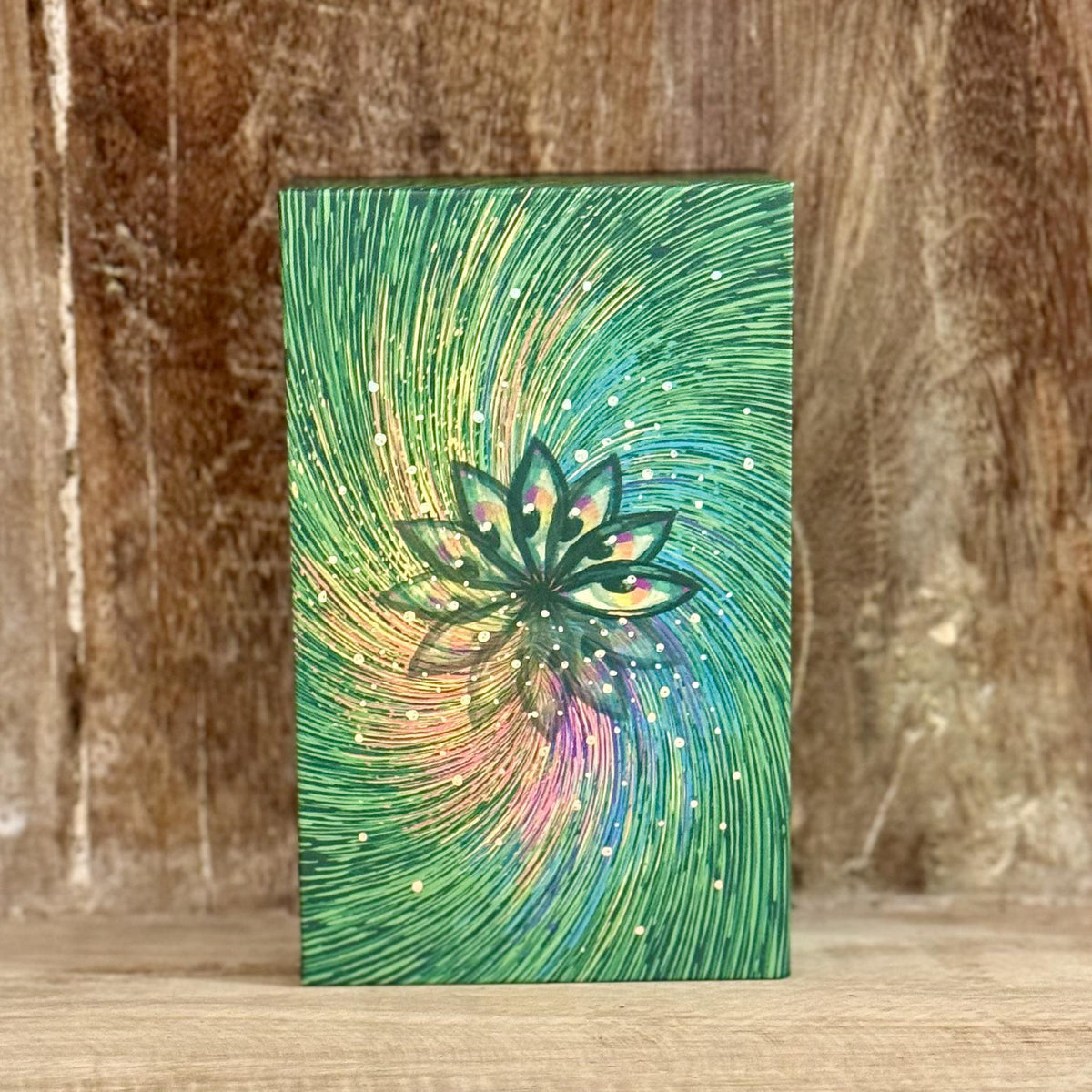 A rectangular object with a cover featuring swirling green, yellow, and pink abstract patterns centered around a lotus flower, reminiscent of the Spellcrafter Cosma Visions Oracle deck.