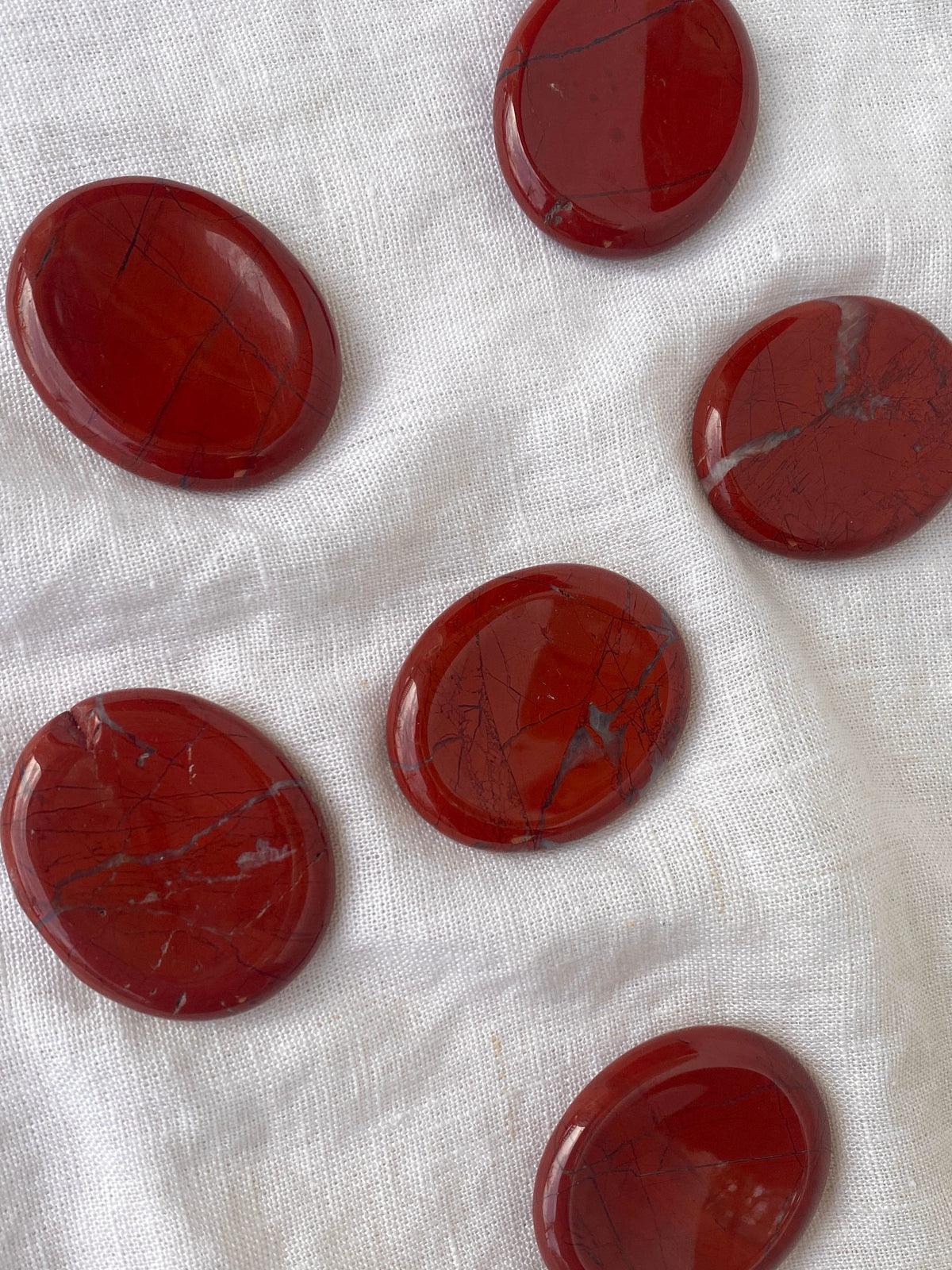 Seven Spellcrafter Red Jasper Calming Stones with dark veins are arranged on a white textured fabric, ideal for Sacral chakra work or as a healing stone display.