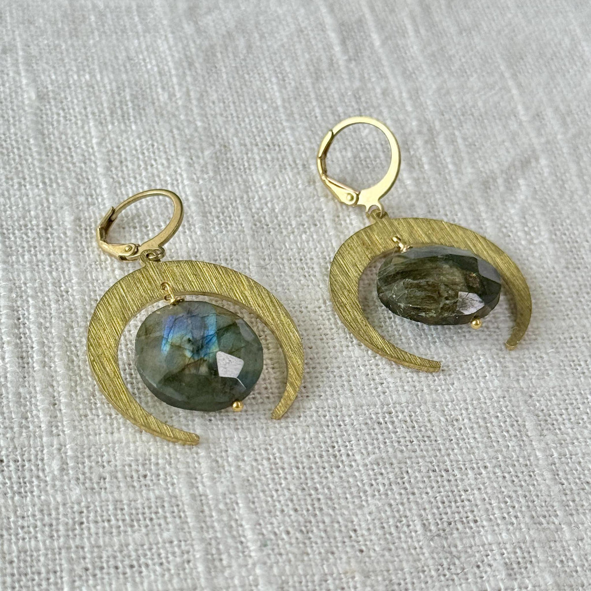 The Spellcrafter Labradorite Lunar Eclipse Earrings feature gold crescent moons with oval, faceted labradorite gemstones at the center, shown on textured white fabric.