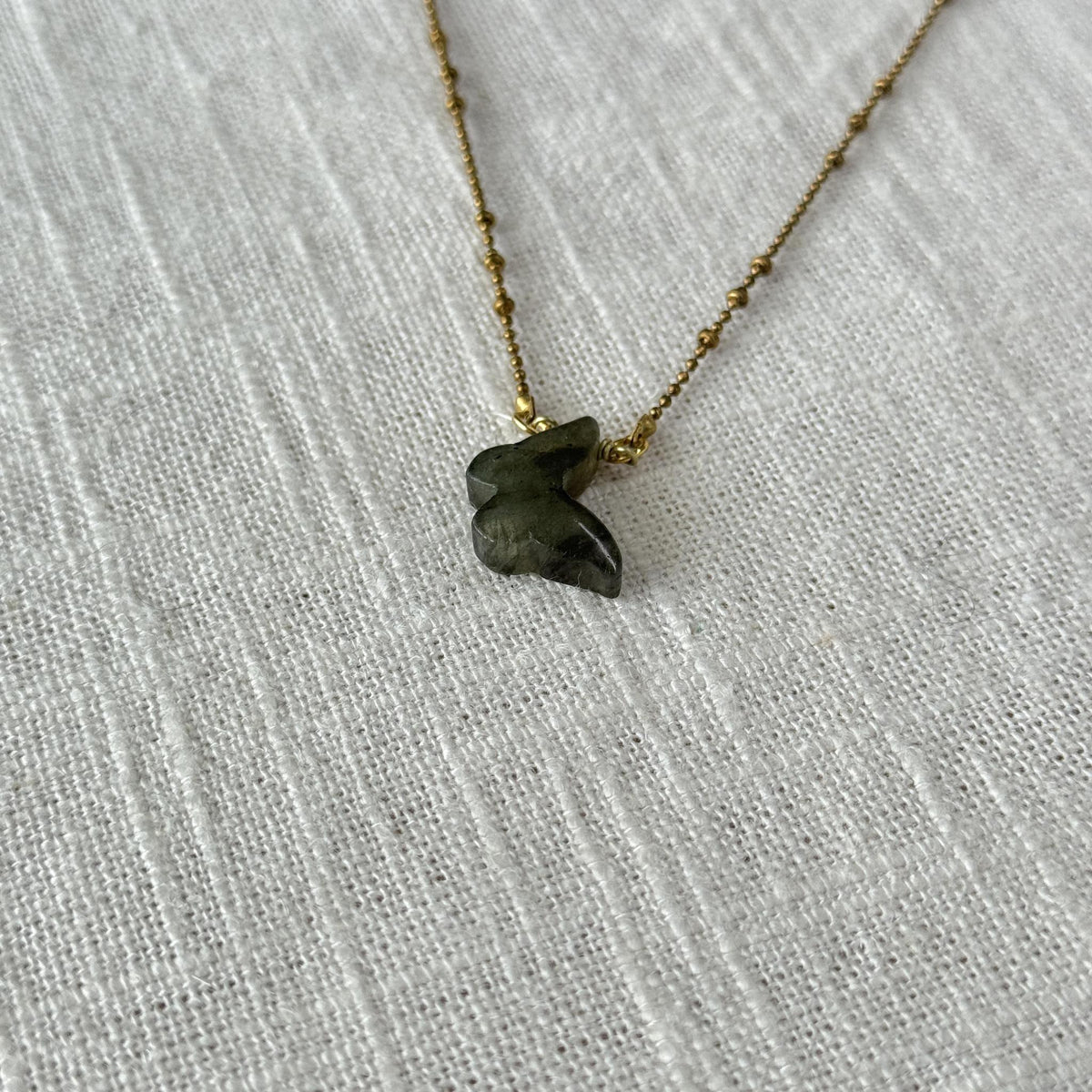 A close-up of the Spellcrafter Labradorite Butterfly Necklace, featuring a small, dark green gemstone pendant on a gold chain, resting on textured white fabric—an elegant and unique jewelry piece.