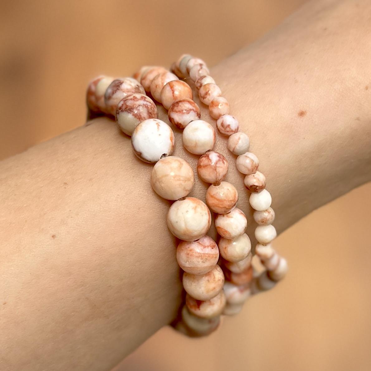 A close-up shows a wrist wearing three Spellcrafter Red Vein Jasper Crystal Bracelets, featuring round beads in cream, peach, and light brown—known as a recovery stone connected to the Root Chakra. Warm tones blur in the background.