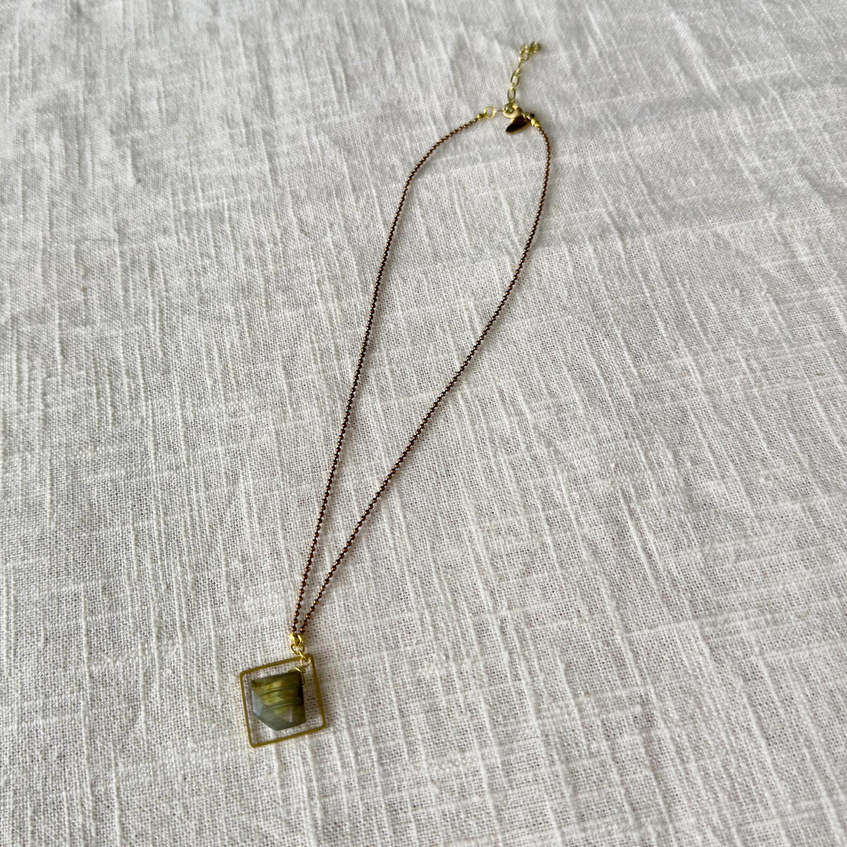 The Spellcrafter Labradorite Framed Diamond Necklace features a fine chain and a square pendant with marbled gray and gold hues, displayed against textured light beige fabric.