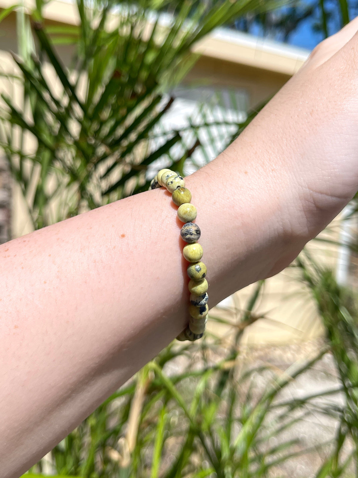 A hand and wrist wearing the Spellcrafter Yellow Agate Bracelet, held up outdoors with palm leaves and a building in the background.