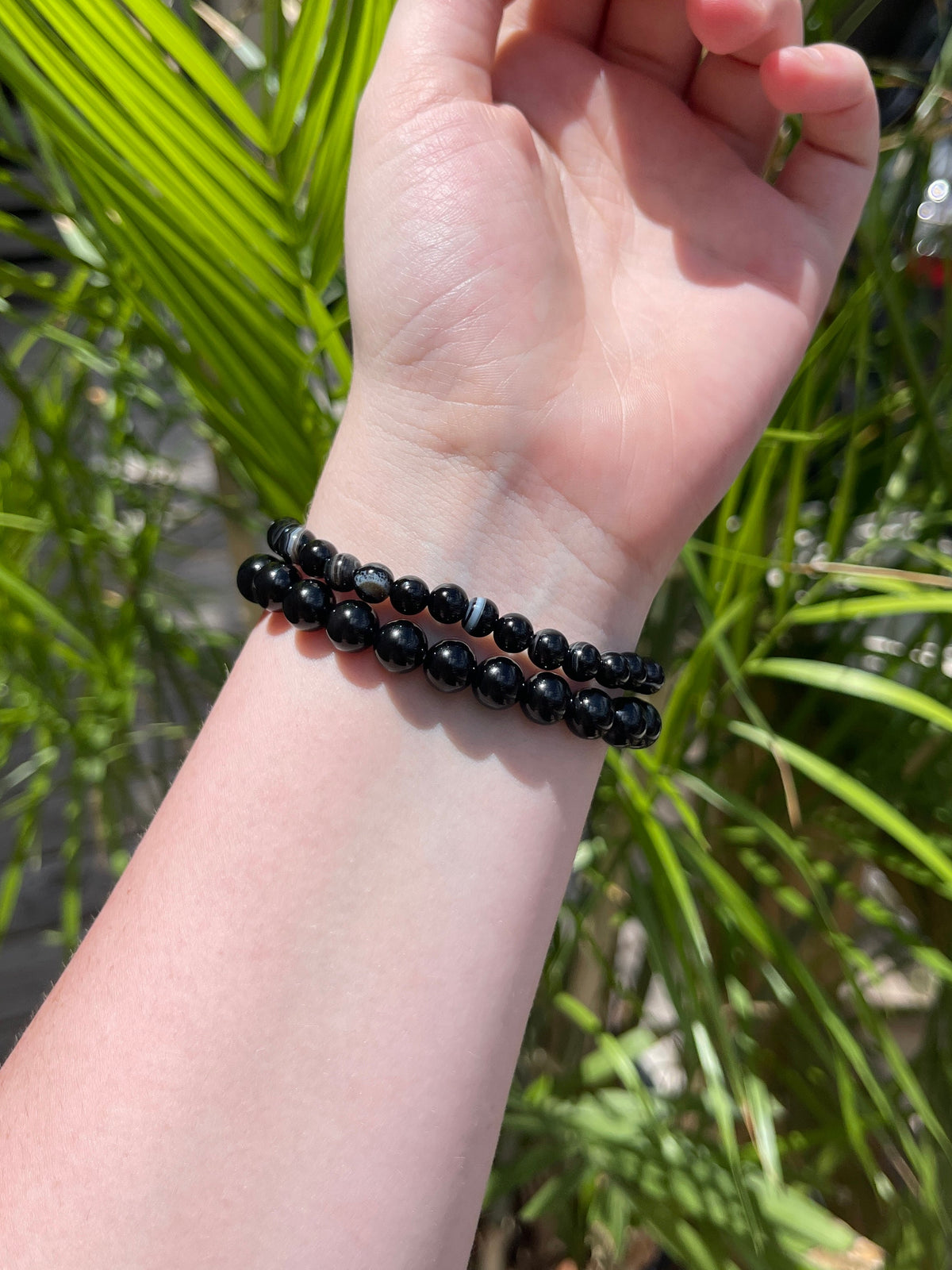 A fair-skinned hand wears two Spellcrafter Black Sardonyx Bracelets, raised in sunlight with blurred green palm leaves in the background.