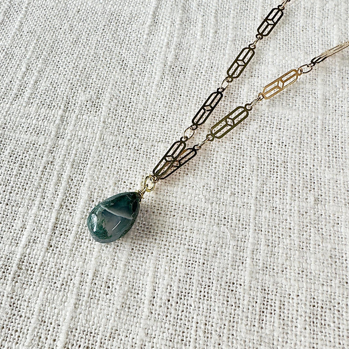 The Spellcrafter Moss Agate Chain Necklace features gold-plated, geometric links and a teardrop-shaped Moss Agate pendant, shown on textured white fabric.