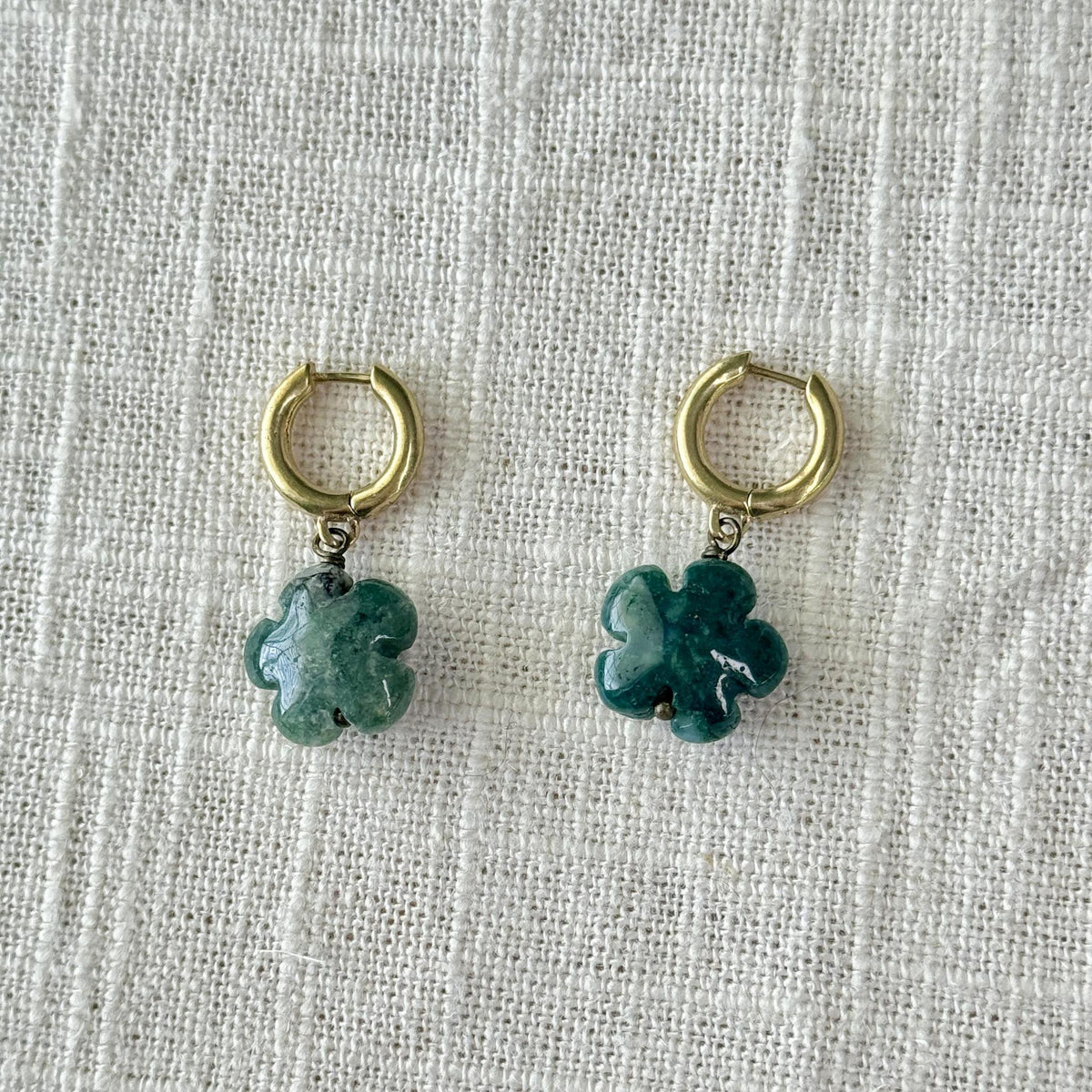 Spellcrafter’s Moss Agate Flower Earrings, gold hoop hypoallergenic earrings with green jade four-leaf clover charms, displayed on a textured white fabric background.