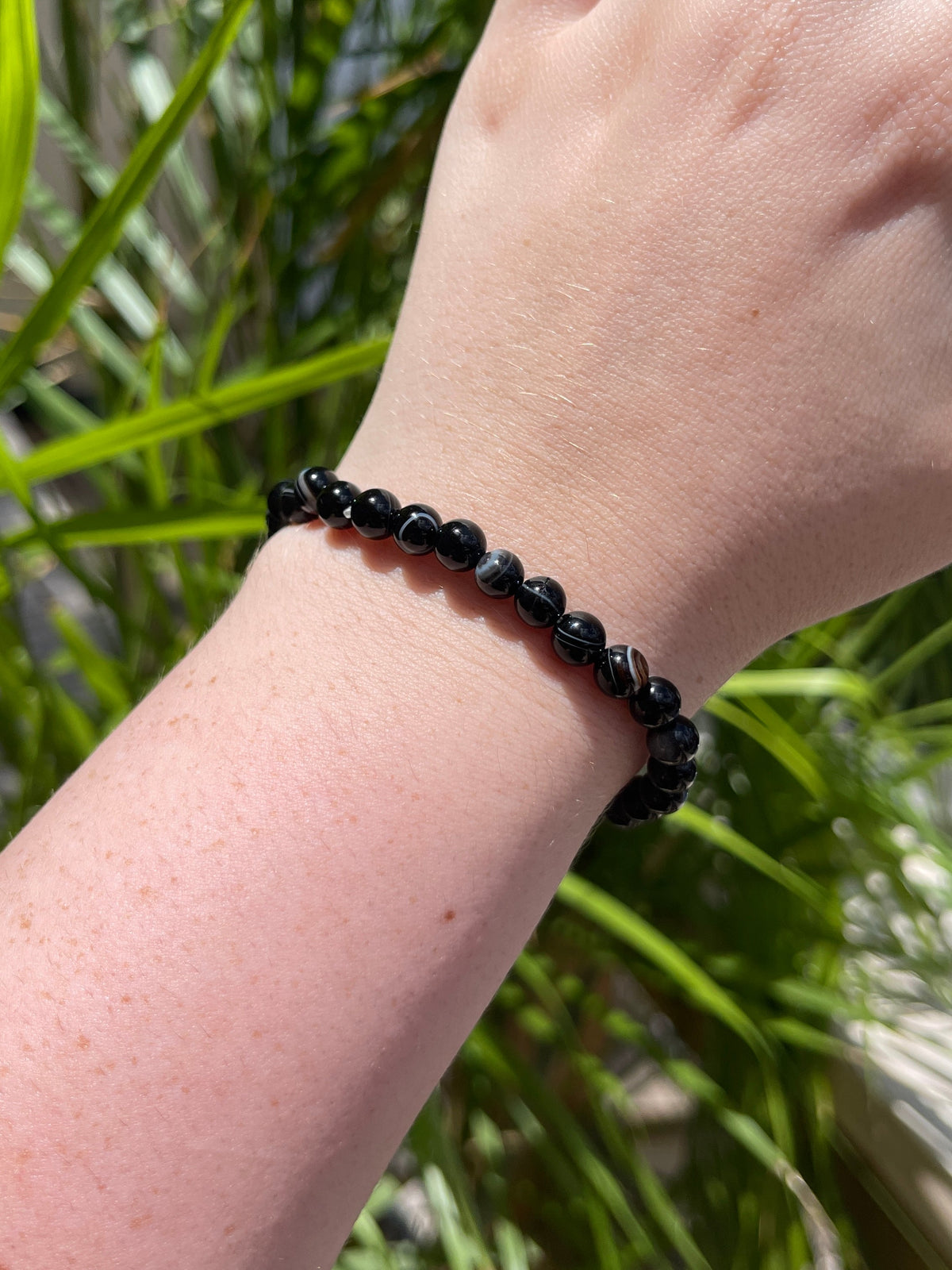 A hand with fair skin wears the Spellcrafter Black Sardonyx Bracelet, featuring small, shiny black beads. Green, leafy plants are blurred in the background while sunlight casts shadows on the wrist.