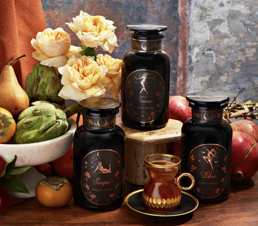 Three black Spellcrafter Zodiac Tea jars with zodiac-themed labels sit by a glass of...