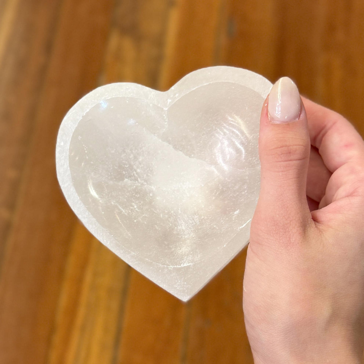 A manicured hand holds the Spellcrafter Selenite Heart Shaped Bowl, a translucent crystal dish symbolizing harmony and cleansing, with a blurred wooden floor in the background.