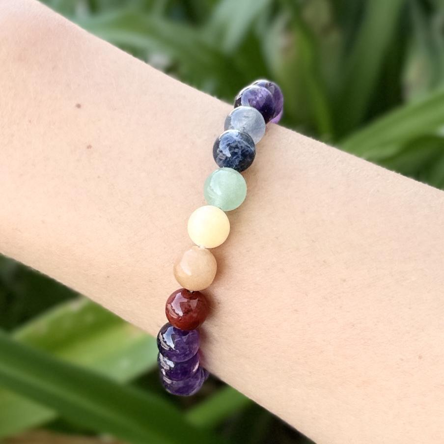 A close-up of the Spellcrafter Chakra Stone Bracelet, featuring multicolored beads on a wrist, set against a softly blurred green outdoor background.