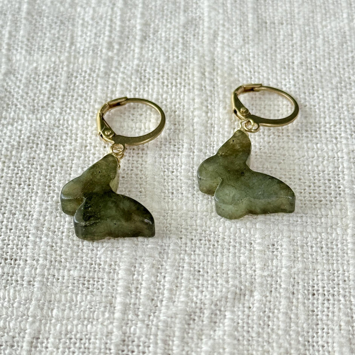 The Spellcrafter Labradorite Butterfly Earrings feature gold hoops with green labradorite butterfly charms, beautifully displayed on a textured white fabric background.