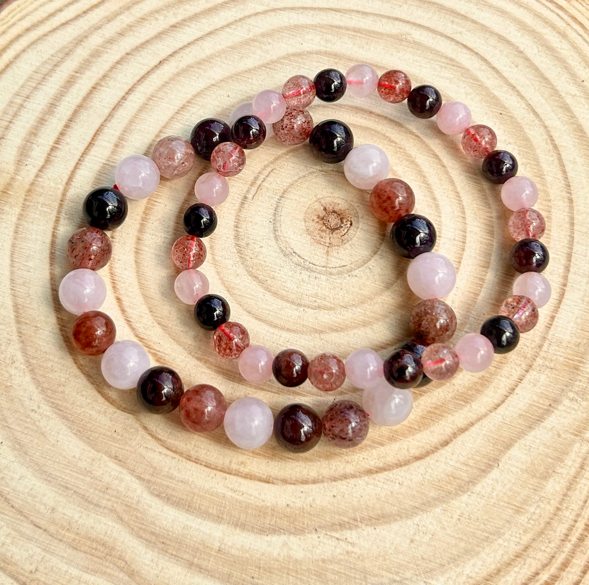 The Spellcrafter Heart Chakra Trio Crystal Bracelet features round pink, red, and maroon beads, reminiscent of rose quartz, and is shown on a circular light wooden surface—perfect for heart chakra support.