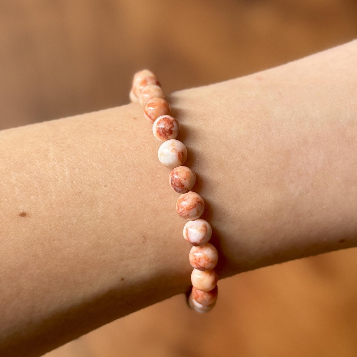 A close-up of a wrist wearing the Spellcrafter Red Vein Jasper Crystal Bracelet—crafted with round, marbled pink and cream beads believed to aid recovery and support the Root Chakra—set against a softly blurred, warm-toned background.