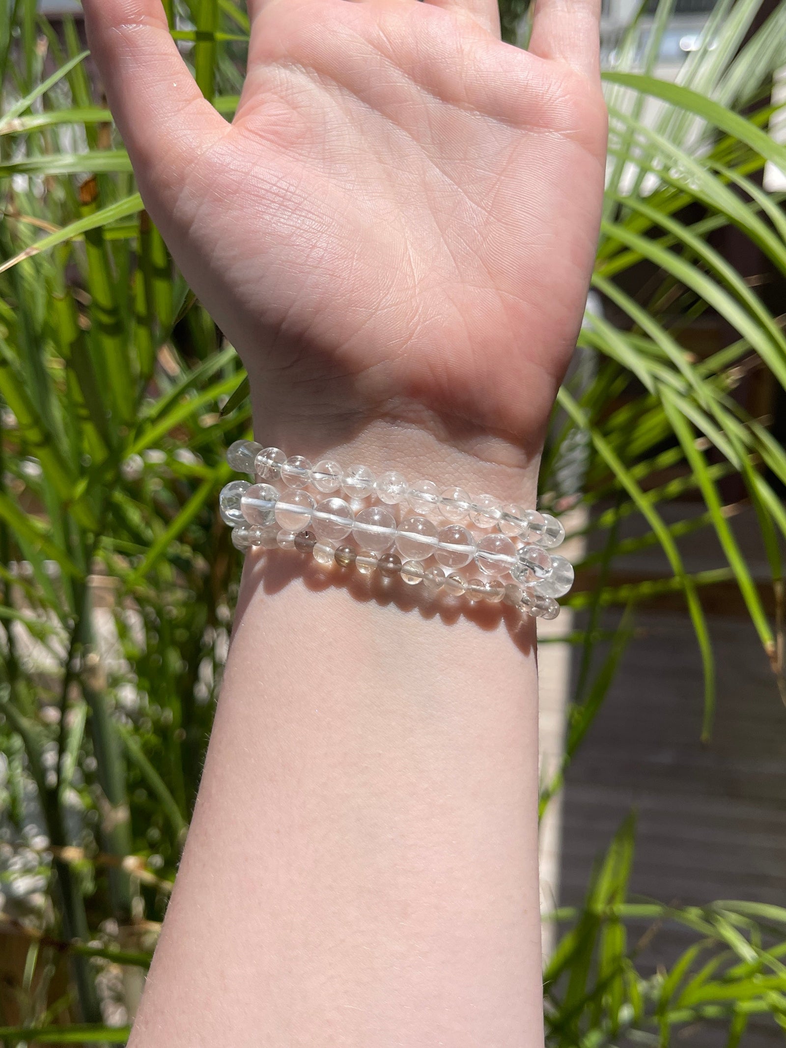 A hand and wrist wearing three Spellcrafter Cracked Clear Quartz Bracelets is shown outdoors...