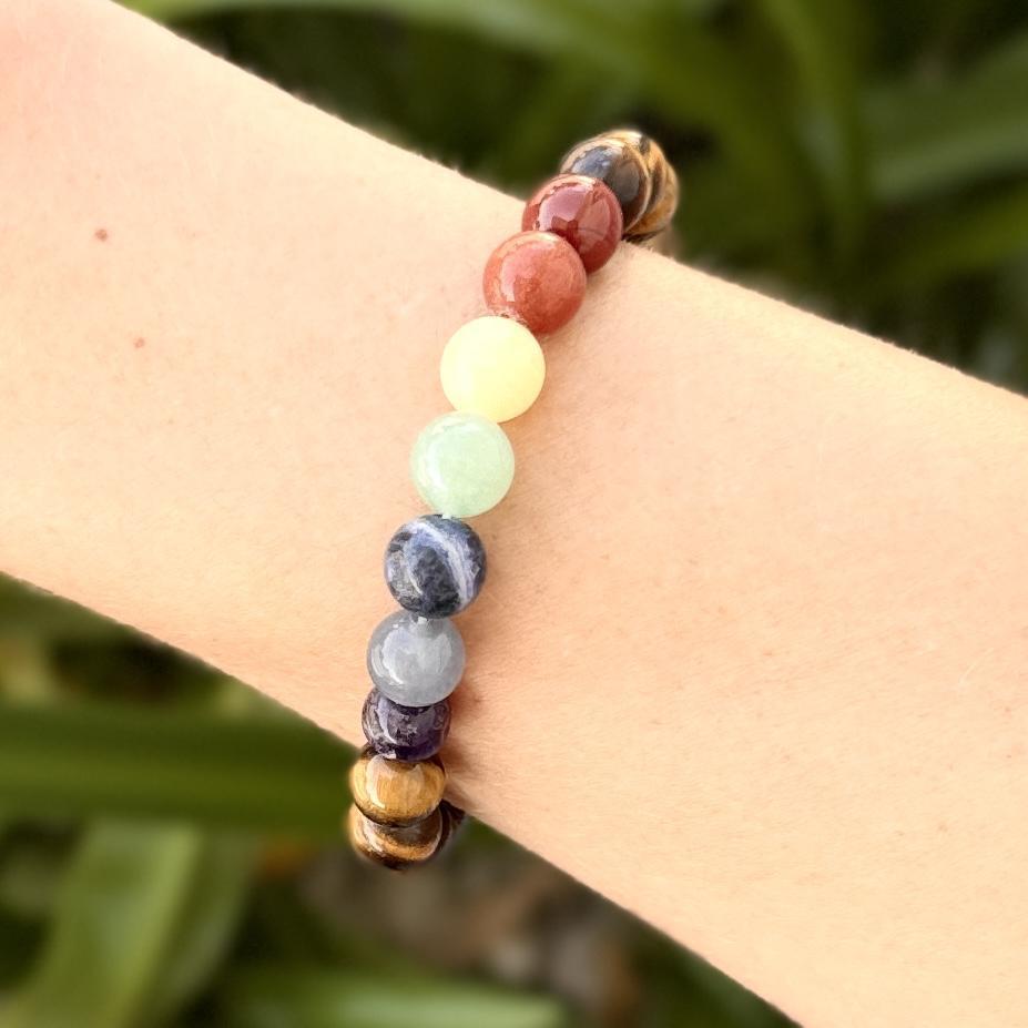 The Spellcrafter Chakra Stone Bracelet, featuring multicolored stones arranged in a rainbow gradient, is worn on a wrist against a softly blurred green background.