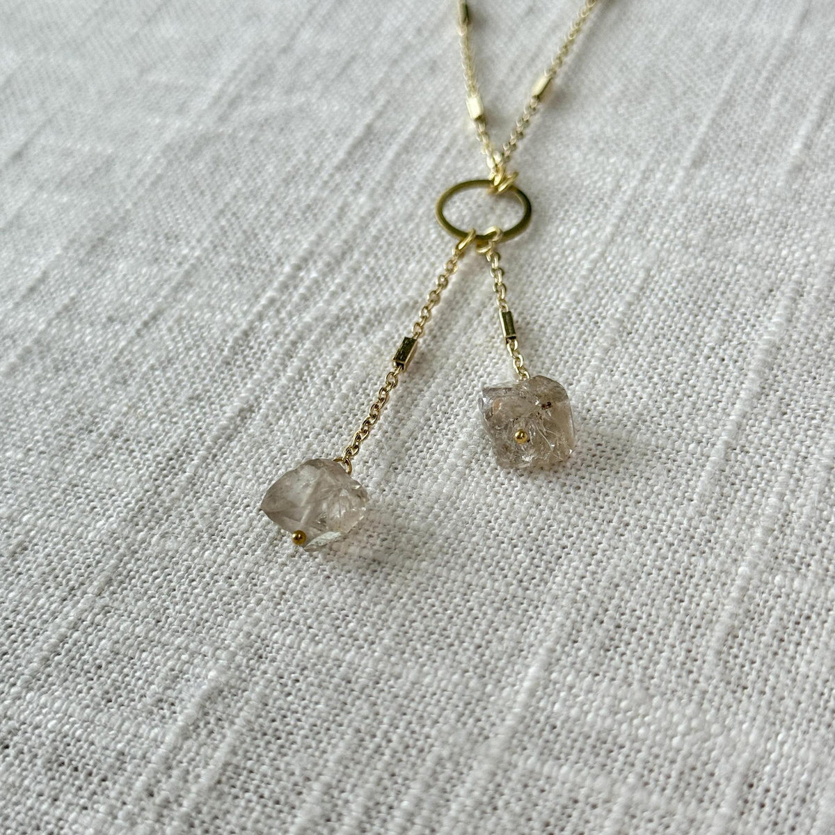 The Spellcrafter Herkimer Diamond Tassel features a delicate gold chain with a circular link and two rough Herkimer Diamond quartz pendants, ideal for meditation, shown on textured white fabric.