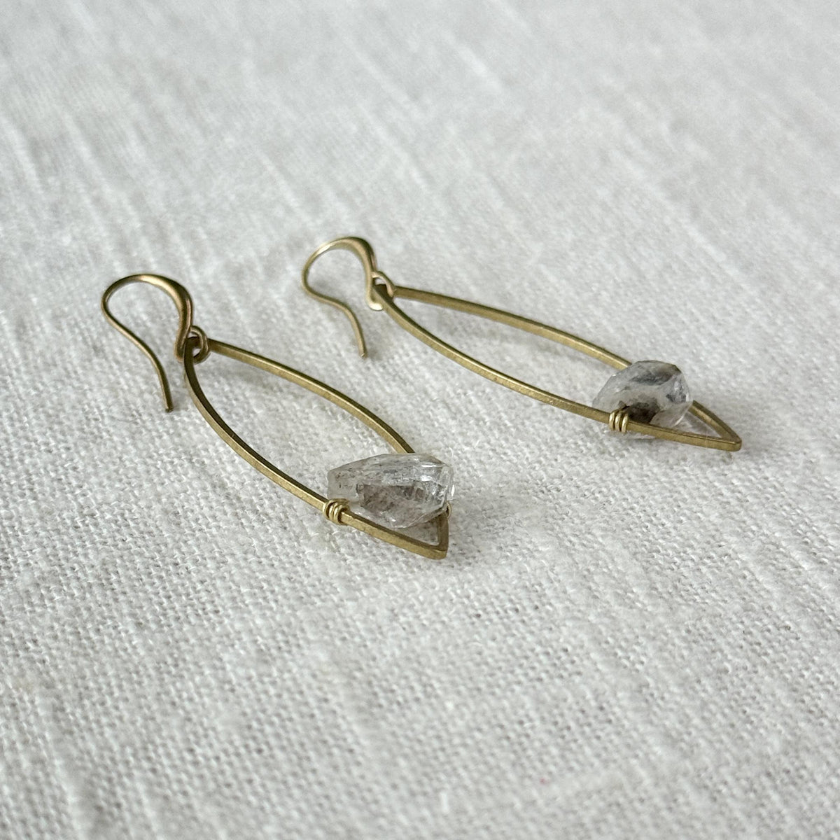 Spellcrafter's Herkimer Floating Marquise Earrings feature elongated gold wire dangles, each with a clear Herkimer Diamond meditation stone at the base, displayed on a textured white fabric background.