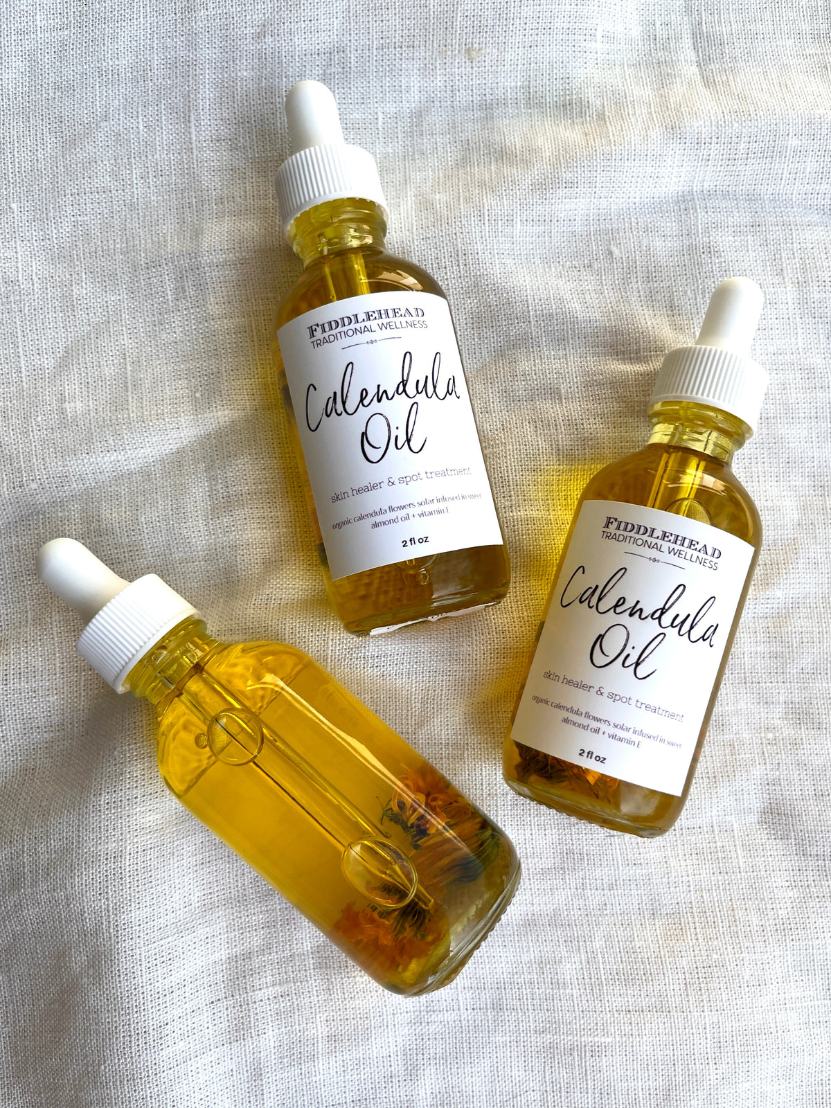 Three glass dropper bottles of Spellcrafter Calendula Oil, each filled with golden herbal-infused oil and calendula petals, sit on textured white cloth—two display the label; one displays the infused flowers.