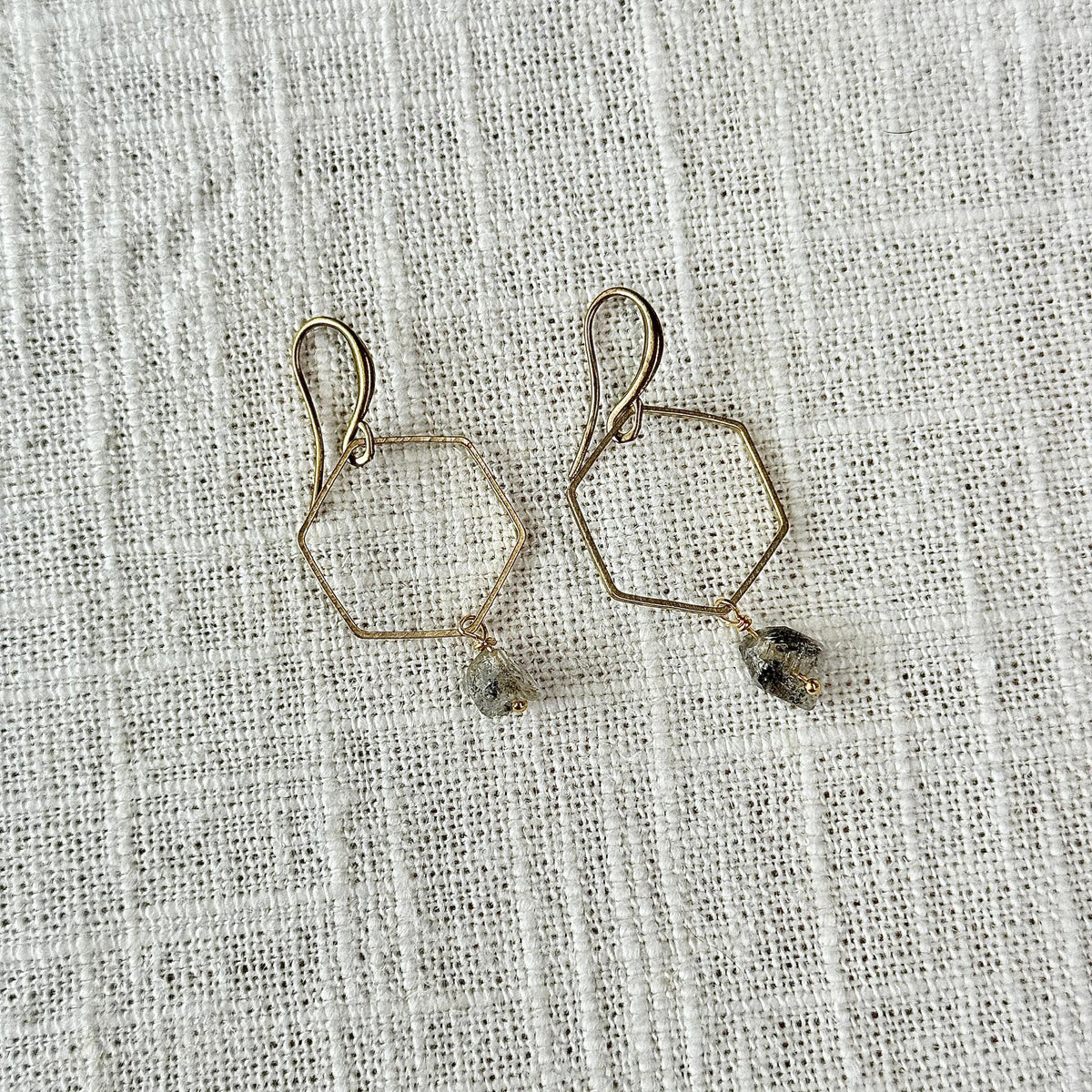 Spellcrafter’s Herkimer Hexagon Earrings feature gold hexagon wire shapes adorned with small faceted Herkimer Diamond beads, shown against a textured white fabric background.