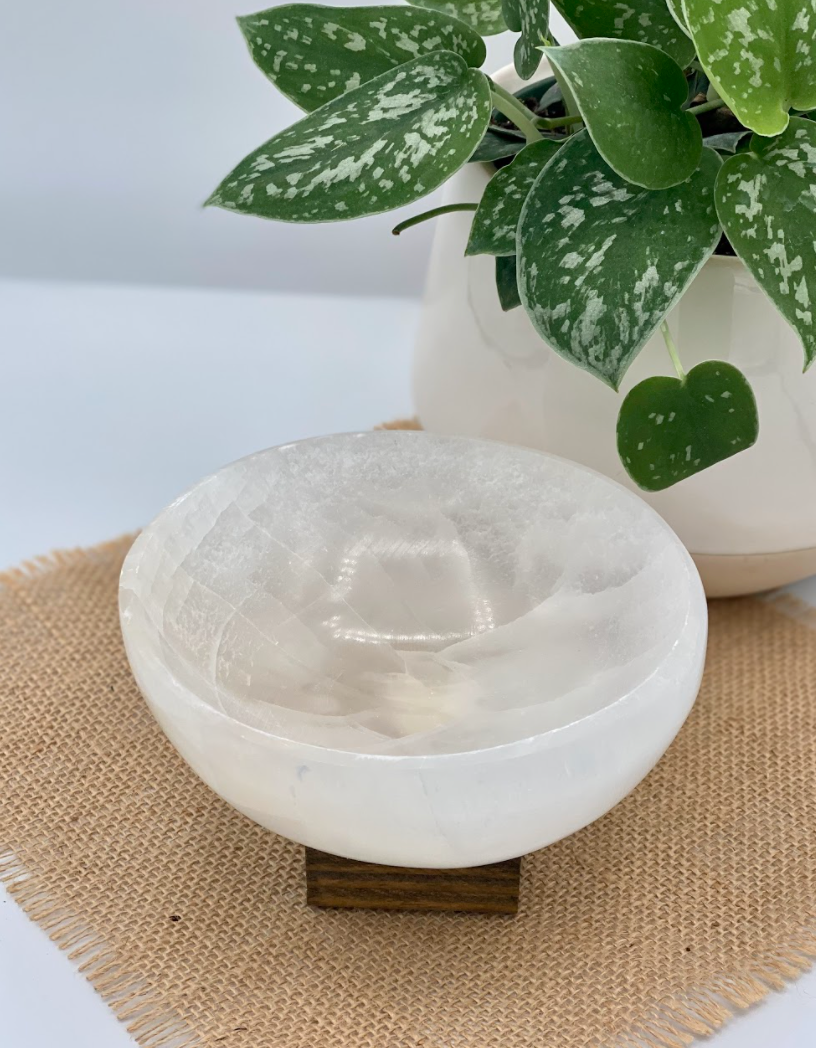 The Spellcrafter Large Selenite Bowl, round and white, rests on a small wooden stand atop a burlap mat, with a leafy potted plant in the background.